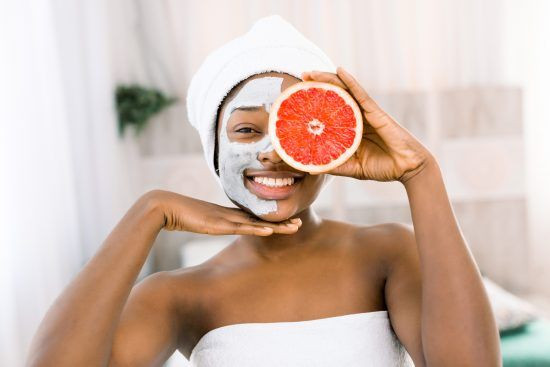 a woman wearing a clay mask holds a grapefruit to her face, playfully covering one eye, suggesting self-care, relaxation, and embracing natural beauty in a moment of personal retreat.