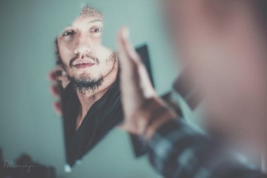 a man in a black shirt thoughtfully looks at his reflection in a shattered mirror, symbolizing broken self-image and the need for positive self-talk.