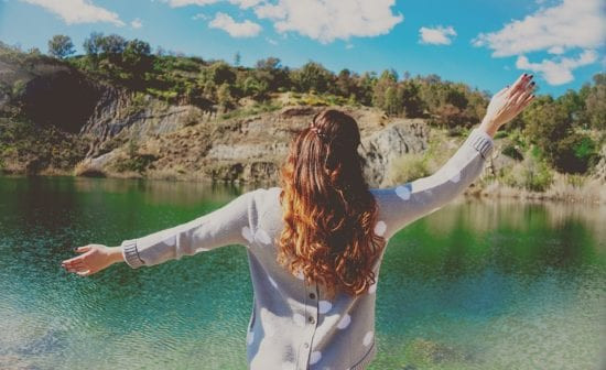 a woman stands with arms outstretched, facing a serene lake at sunset, embodying a sense of freedom, peace, and gratitude for the beauty and abundance in life.