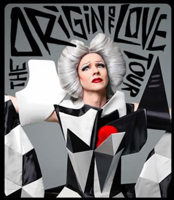 John Cameron Mitchell performing on stage during The Origin of Love concert tour, wearing a black, white, gray, and red costume, and Hedwig's iconic wig, surrounded by a cheering audience.